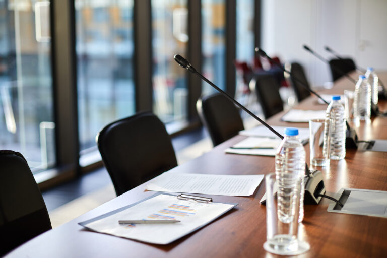 Business documents, plastic bottles of water, glasses and microphones along table and chairs near by in conference hall